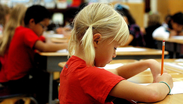 Young girl in a red shirt and pigtails sits at her desk in a full classroom.