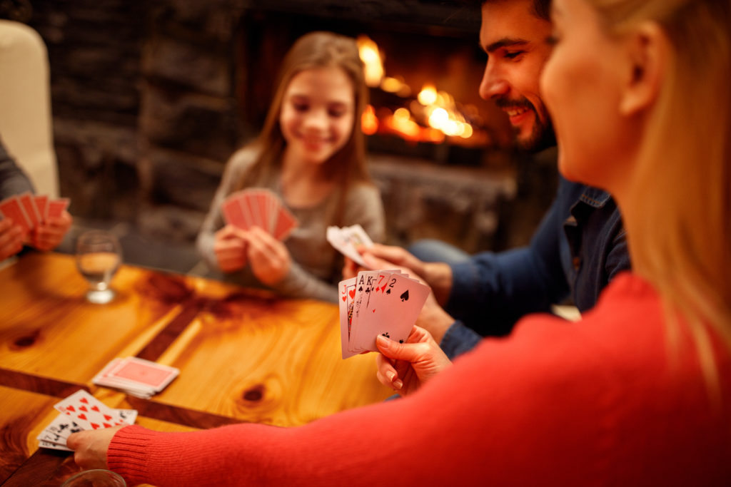Family sits around the table playing cards, with a fire in a fire place behind them.