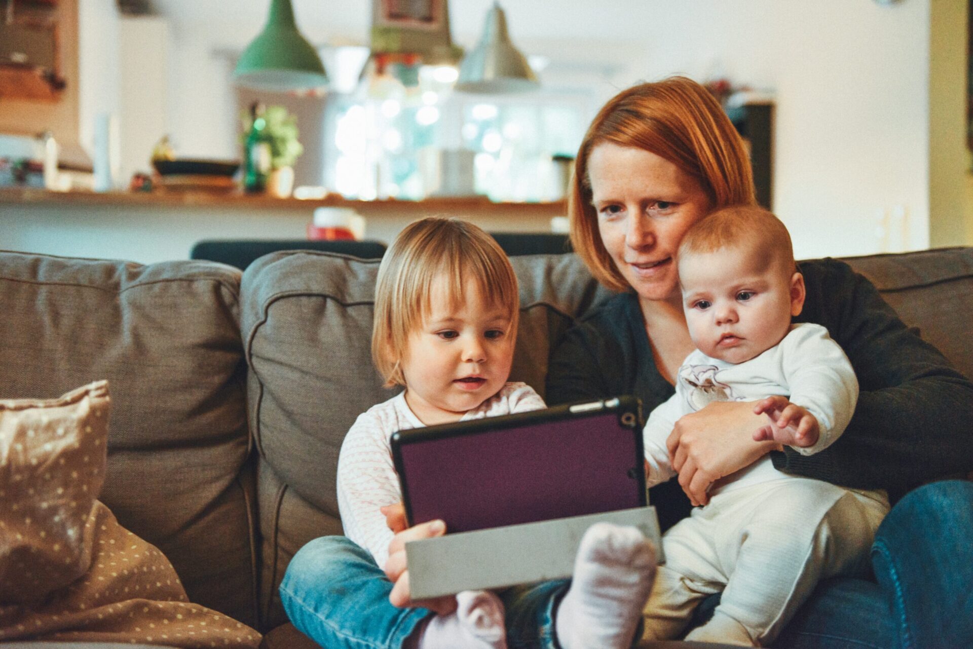 A mother sits on the couch with her toddler, baby, and iPad.