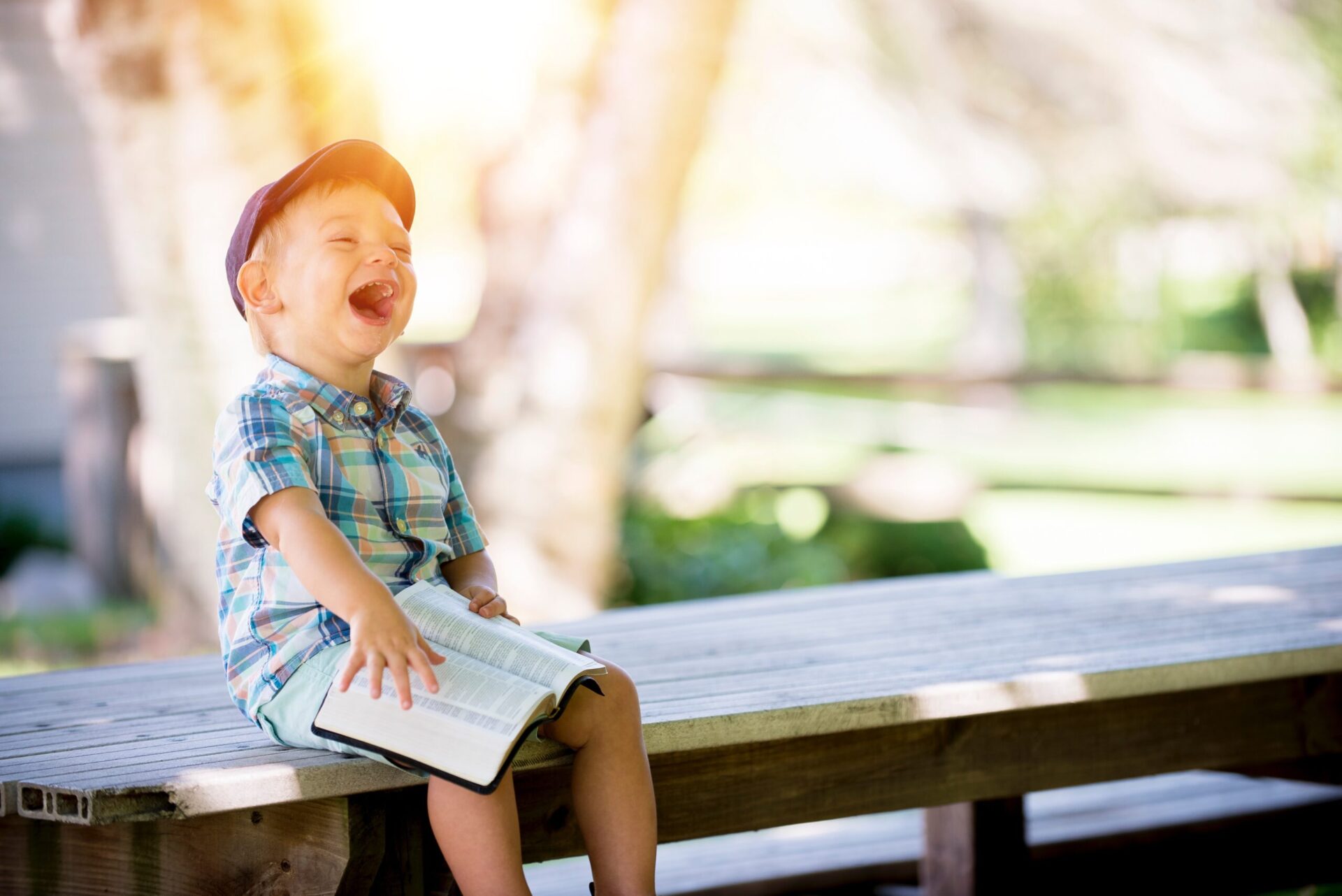 A laughing young boy sits on an outdoor bench with an open book in his hand.