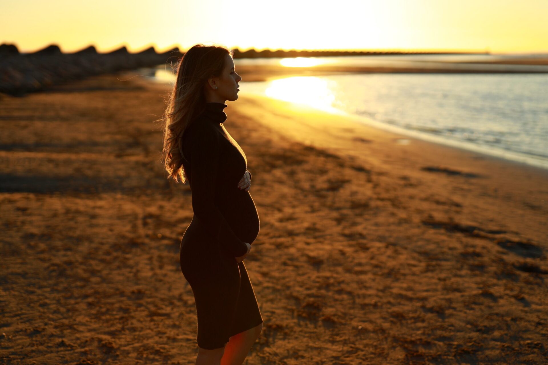 Pregnant woman standing on the beach looks out to the water.