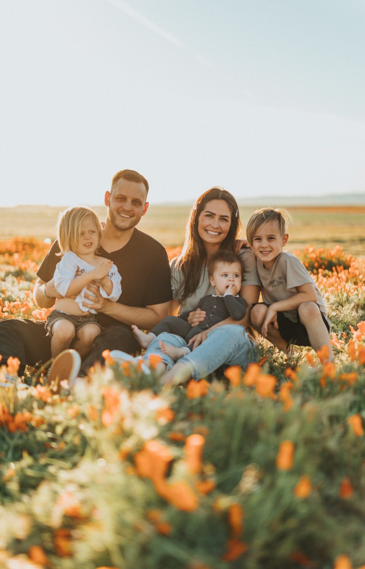 Smiling family of a mother, father and three young children sit in a green field dotted with orange flowers.