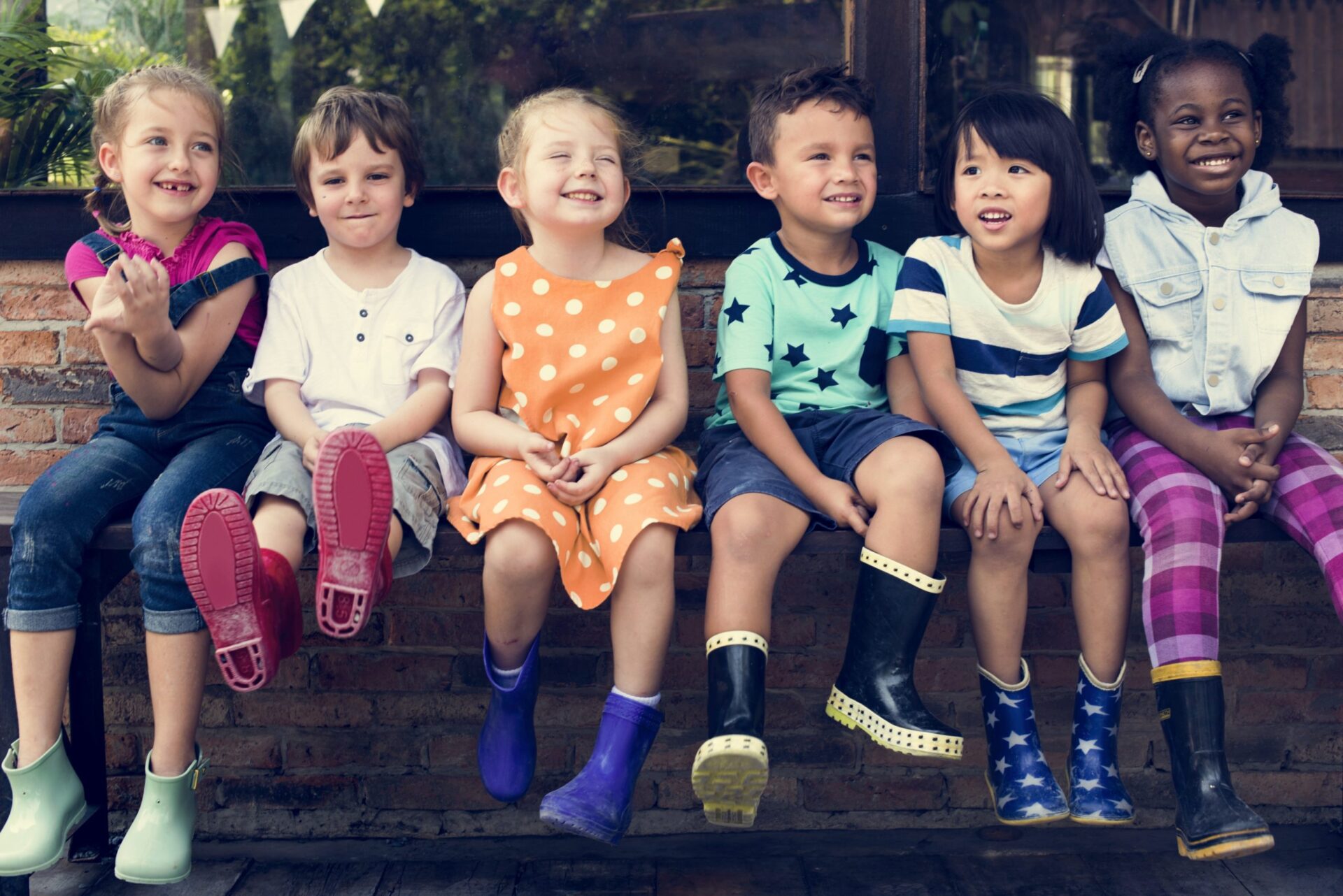 A group of six children, all wearing rainboots, sit outside on a bench.