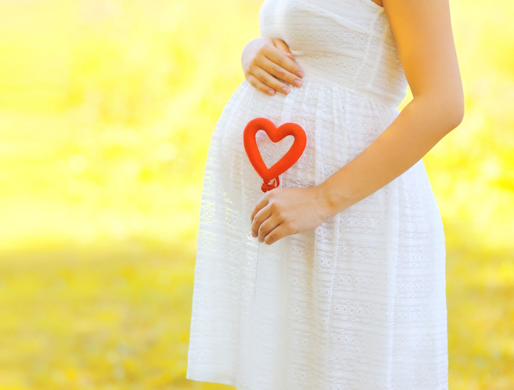 Pregnant woman wearing a white dress has her right hand on her belly and her left hand holding a red heart decoration.