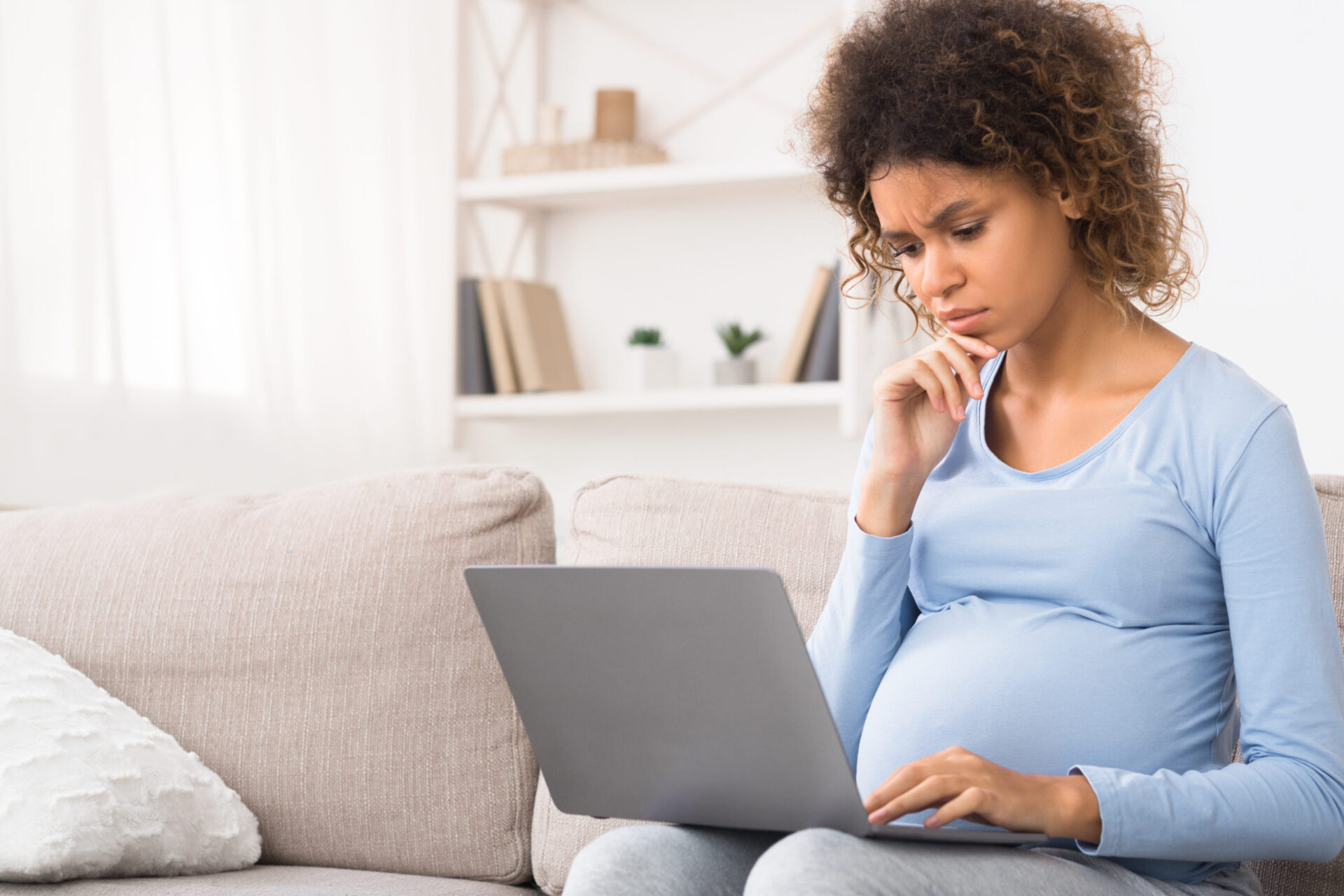 a pregnant woman sits on a couch looking at a laptop with a concerned look