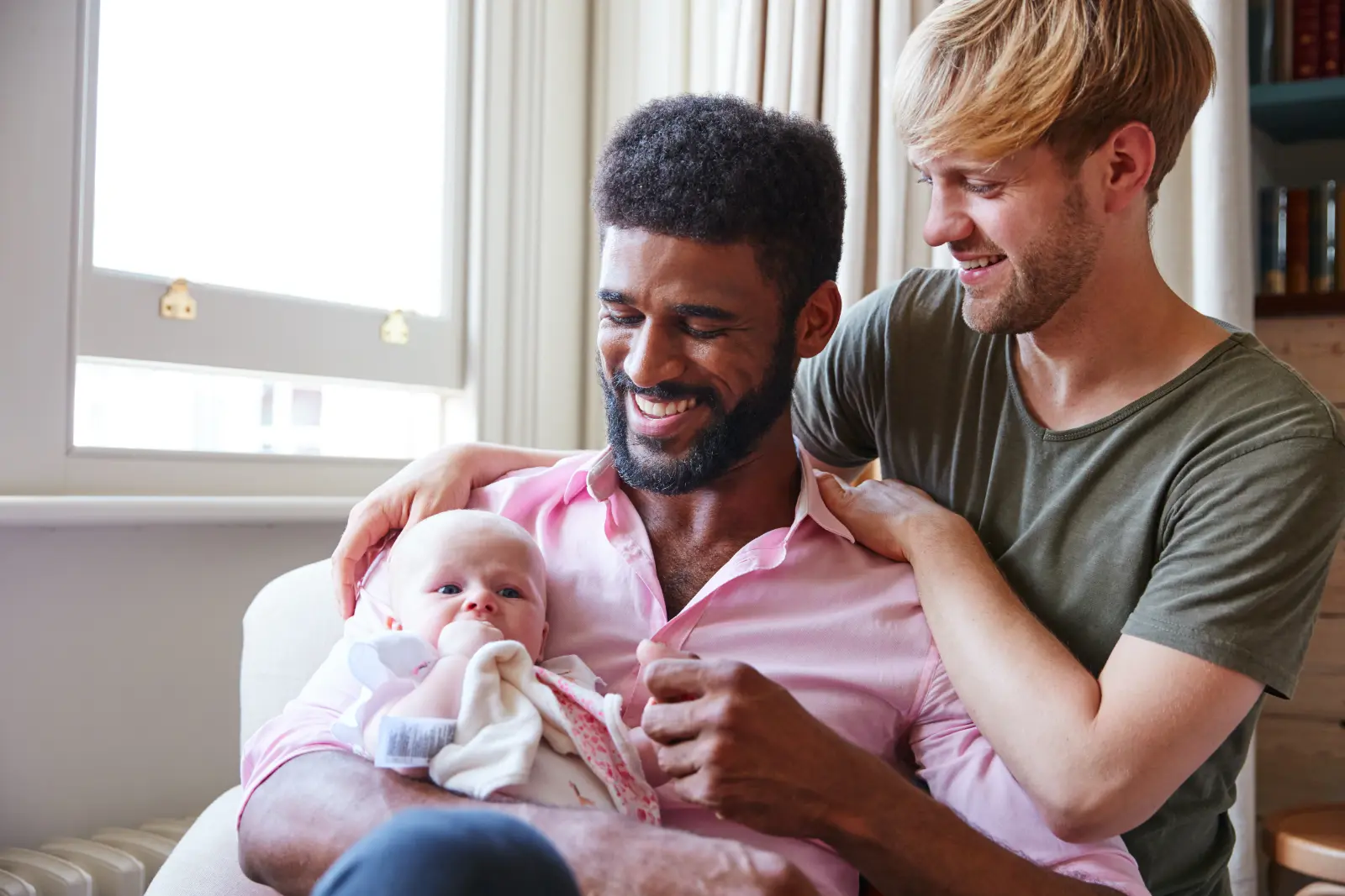 Loving Male Same Sex Couple Cuddling Baby Daughter On Sofa At Home Together. Open Adoption